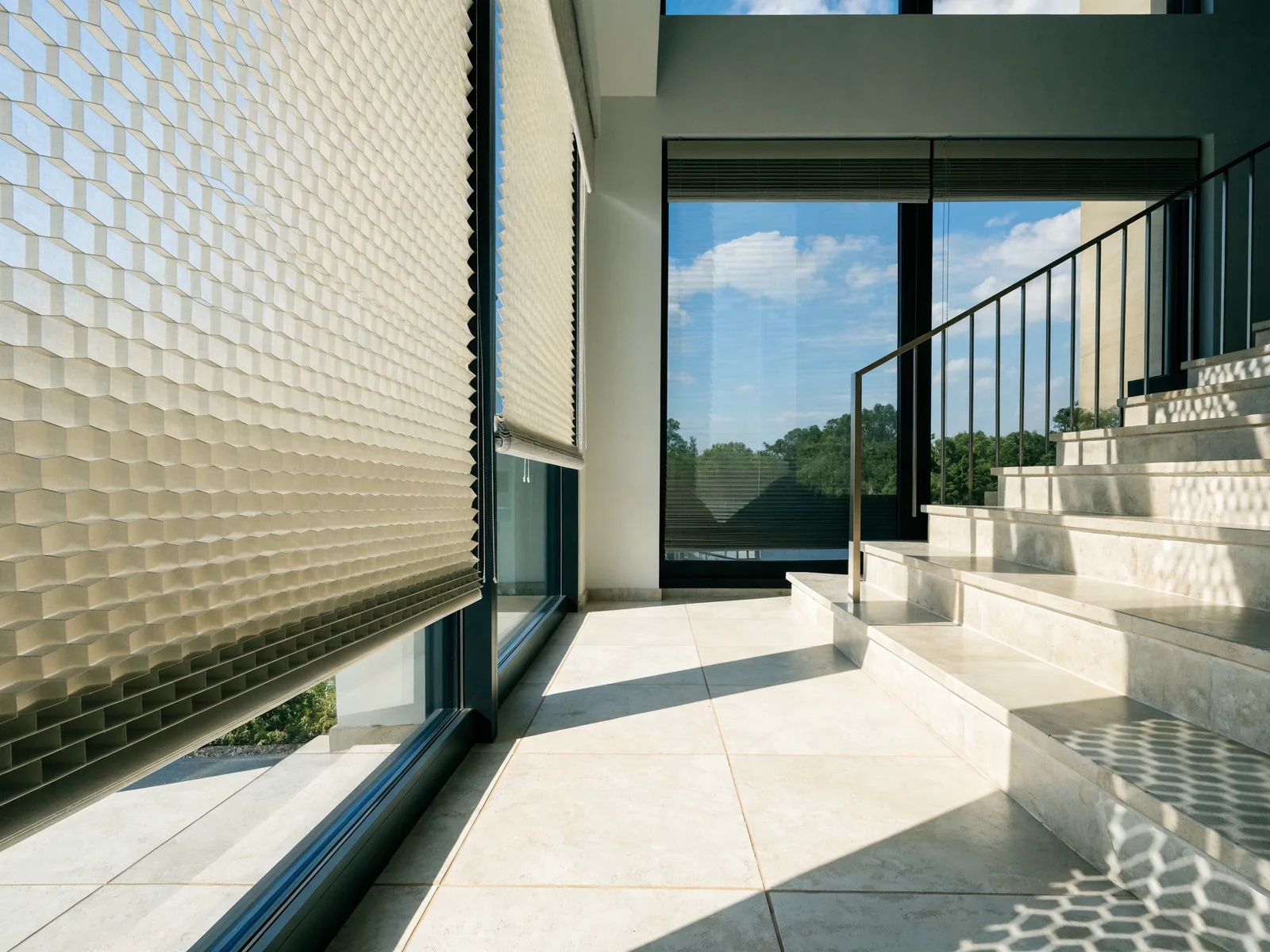 A cellular honeycomb blind, its concertina of hexagonal air cells clearly visible edge-on between two smooth fabric faces, half lowered over the tall glass of a double-volume entrance hall in a modern Midstream estate home, bright Highveld daylight across a pale tiled floor and staircase